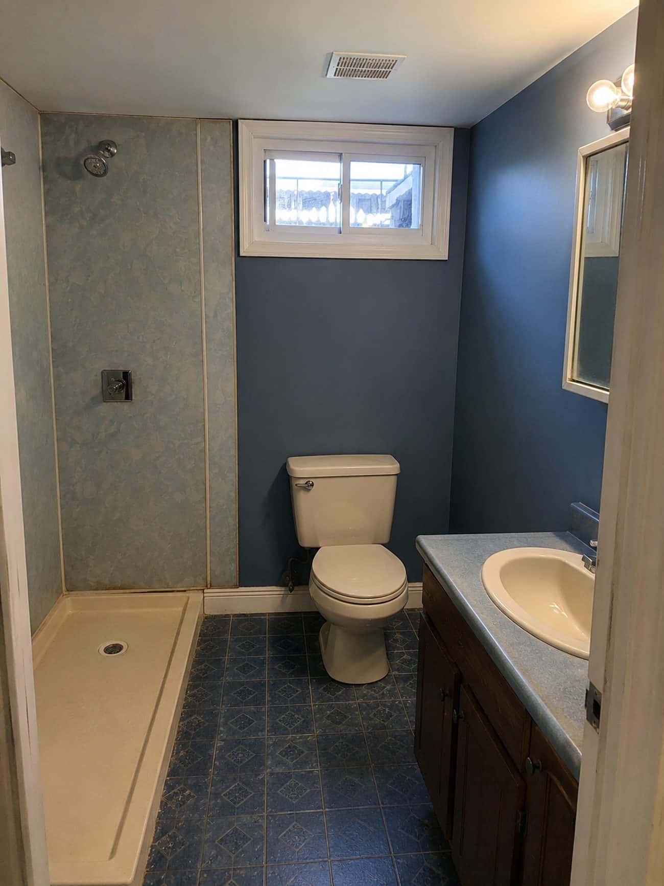 Original basement bathroom in a Gwynn Oak home with a fiberglass shower pan, blue vinyl floor and dated vanity before remodeling