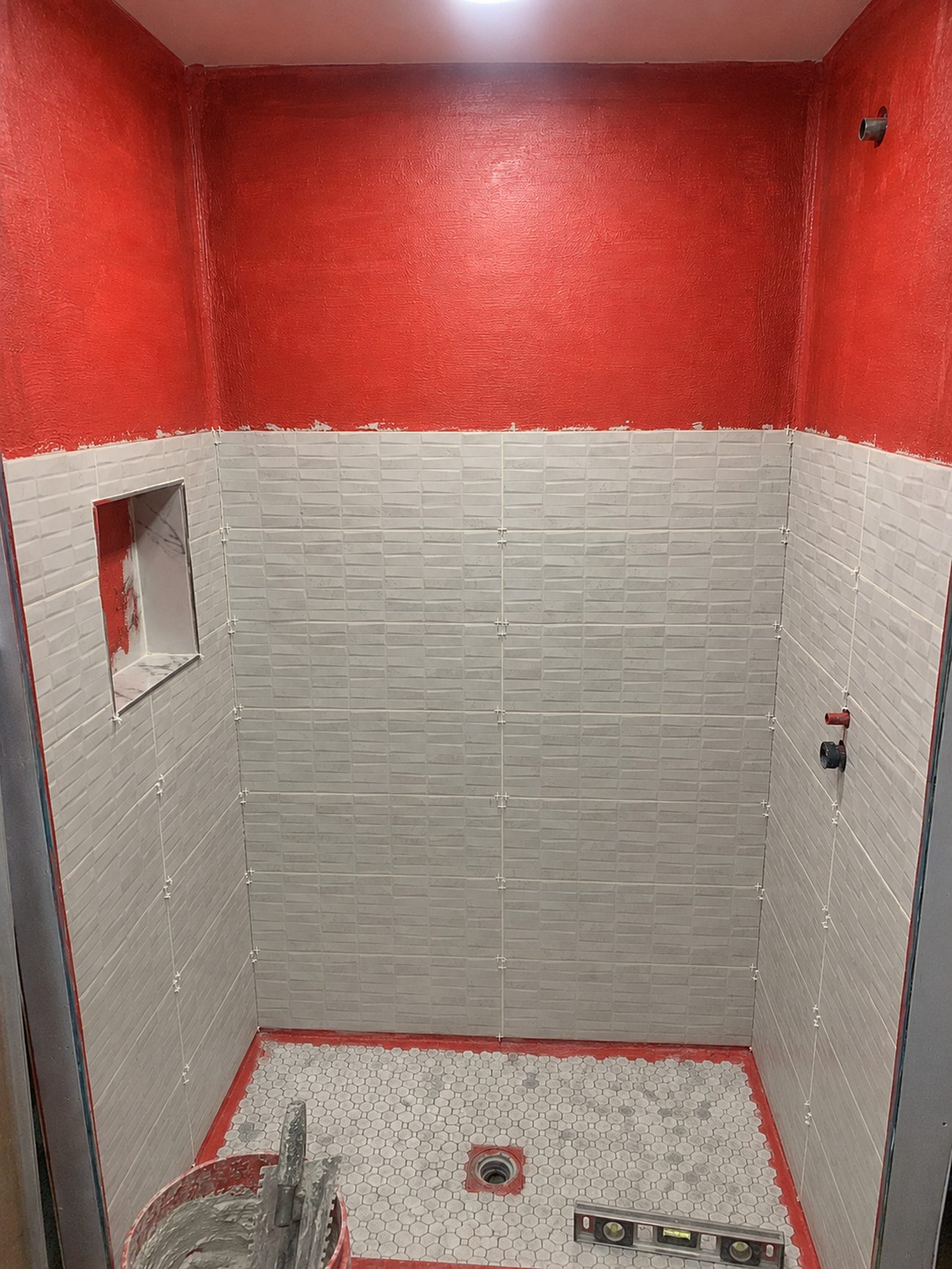 Wall tile installed over red waterproofing membrane in a basement shower, with hex mosaic floor